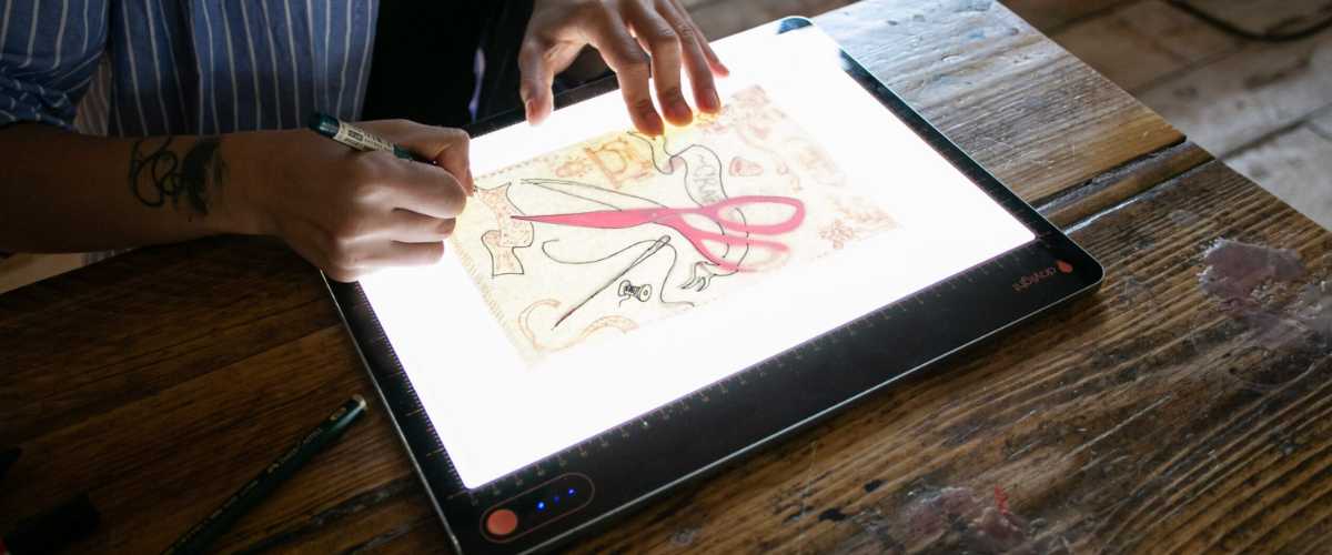 Close-up lifestyle photo of an artist's tattooed hands tracing a colourful illustration featuring scissors and decorative motifs on a glowing Daylight LED light pad placed on a rustic wooden table.