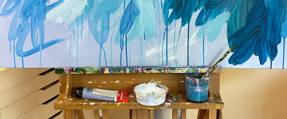 Artist's workspace showing a large blue abstract canvas in progress with a paint-splattered palette and brushes