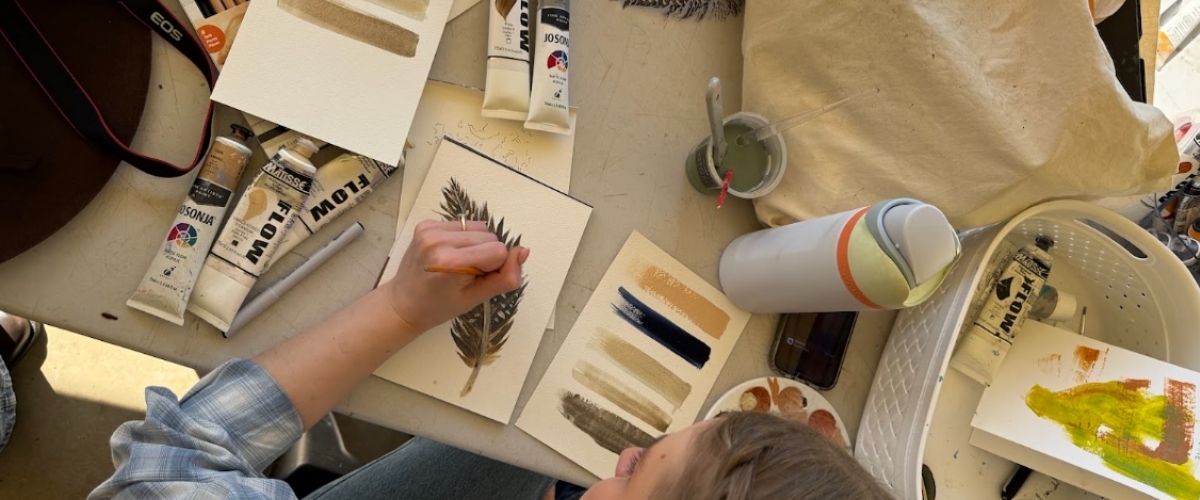 Artist painting a native feather study with Jo Sonja and Flow paint tubes on a studio table